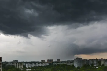 Dark storm clouds loom over a city skyline, with apartment blocks and trees below at dusk/light clouded horizon.