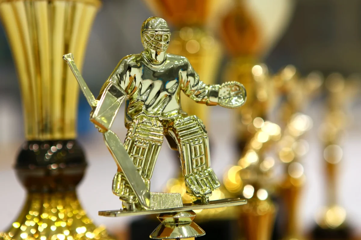 Zlatá trofejní socha hokejisty na reflexním podstavci s dalšími trofejemi rozmazanými v pozadí.