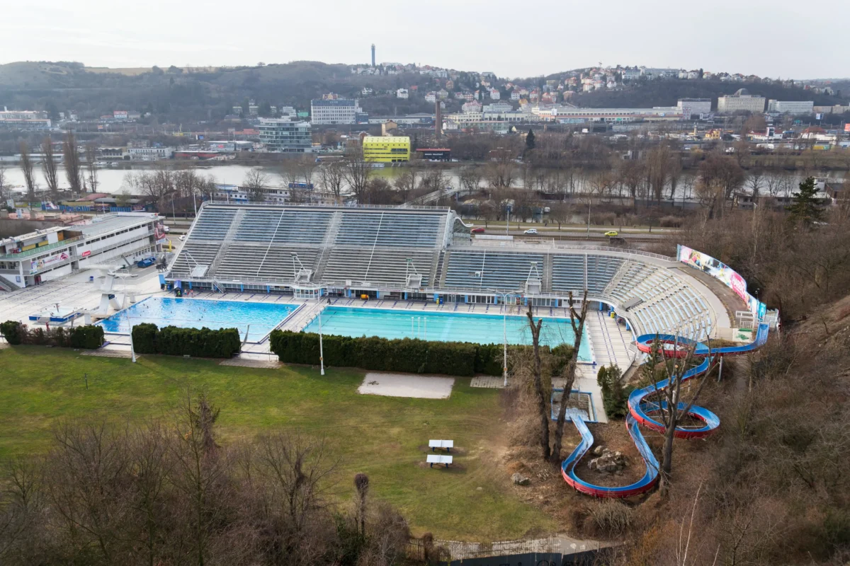 shutterstock_1338261707 1 | TVGURU.cz Aerial view of a stadium with a large pool and winding blue slides in the foreground, city on a hill across a river in the background, trees bare in winter.