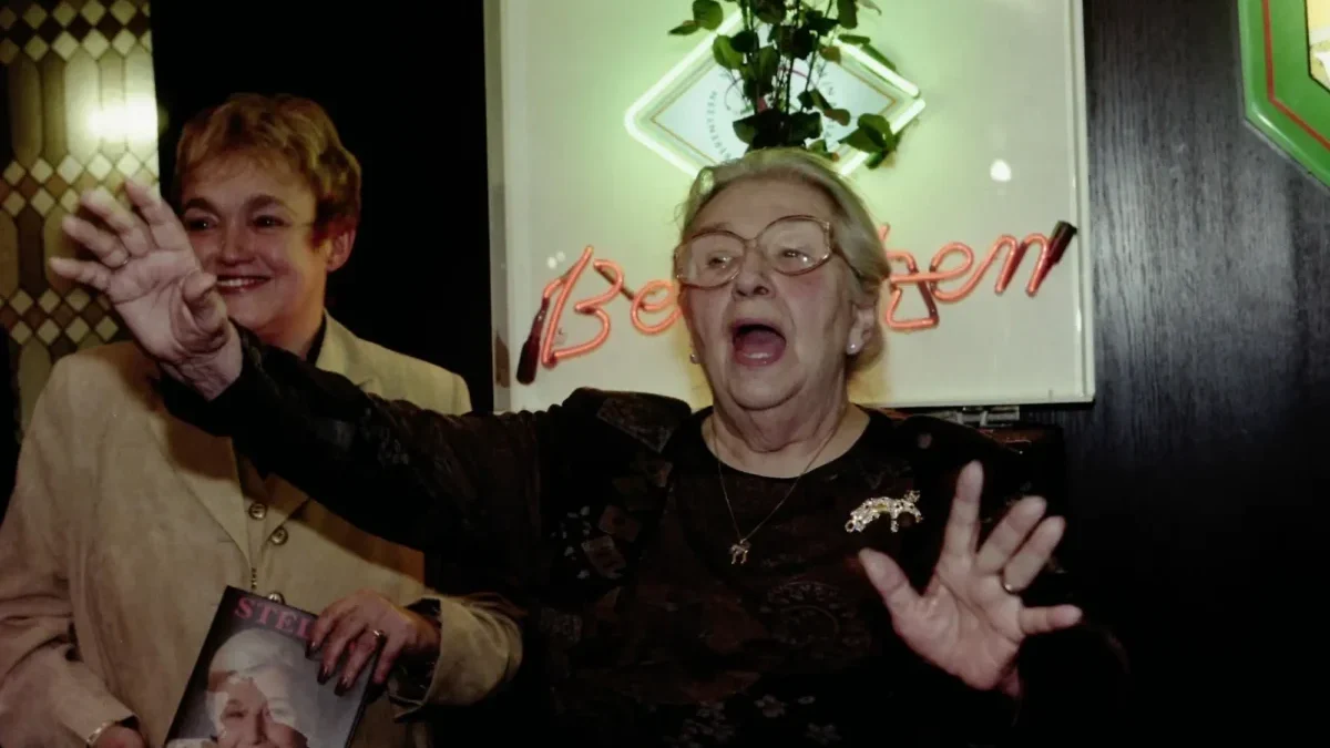 Two smiling older women at a celebration; one waves toward the camera while the other wears glasses and holds up a magazine with a portrait on the cover in front of a neon sign.
