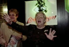 Two smiling older women at a celebration; one waves toward the camera while the other wears glasses and holds up a magazine with a portrait on the cover in front of a neon sign.