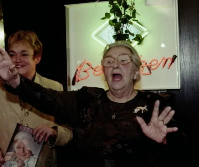 Two smiling older women at a celebration; one waves toward the camera while the other wears glasses and holds up a magazine with a portrait on the cover in front of a neon sign.