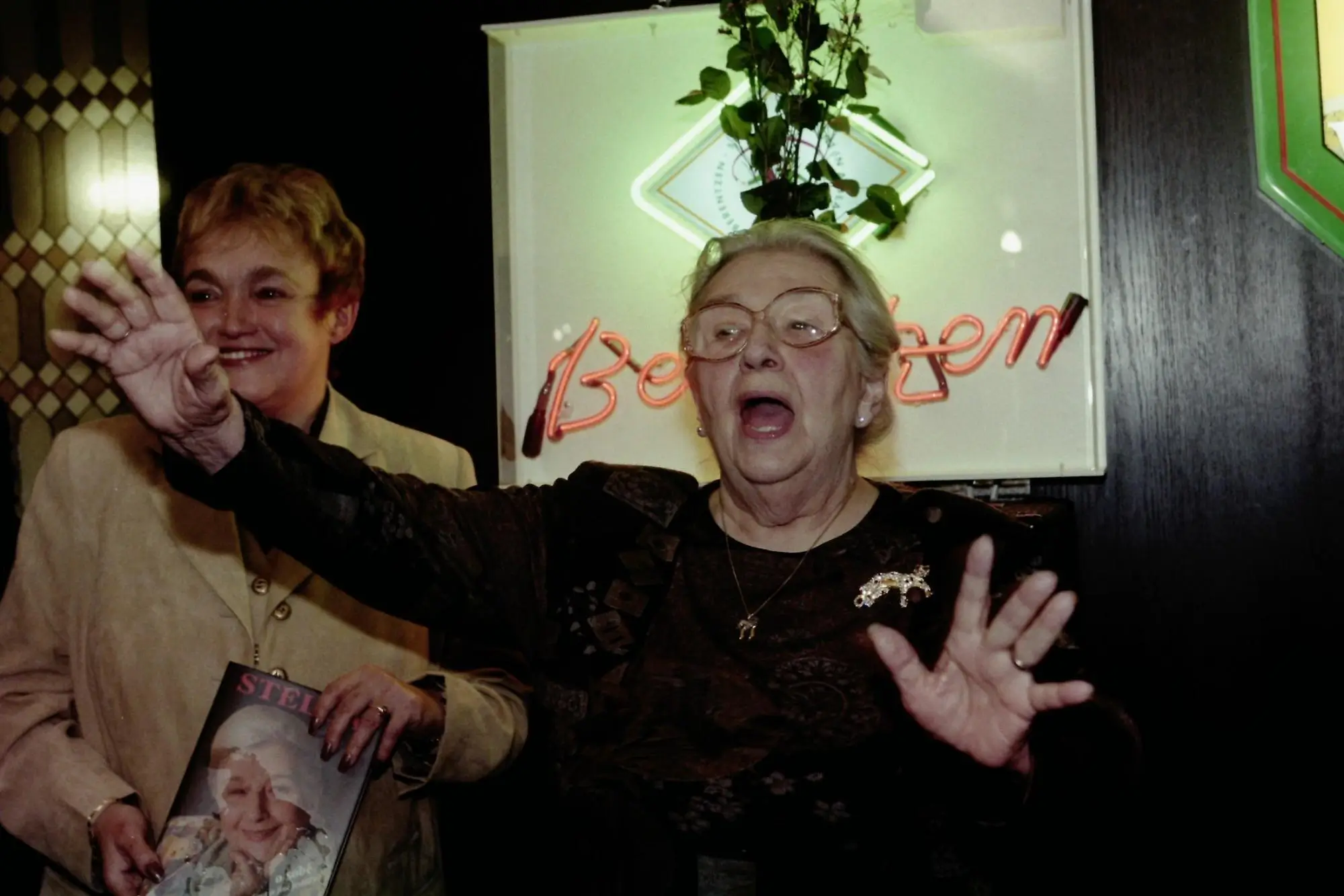 Two smiling older women at a celebration; one waves toward the camera while the other wears glasses and holds up a magazine with a portrait on the cover in front of a neon sign.