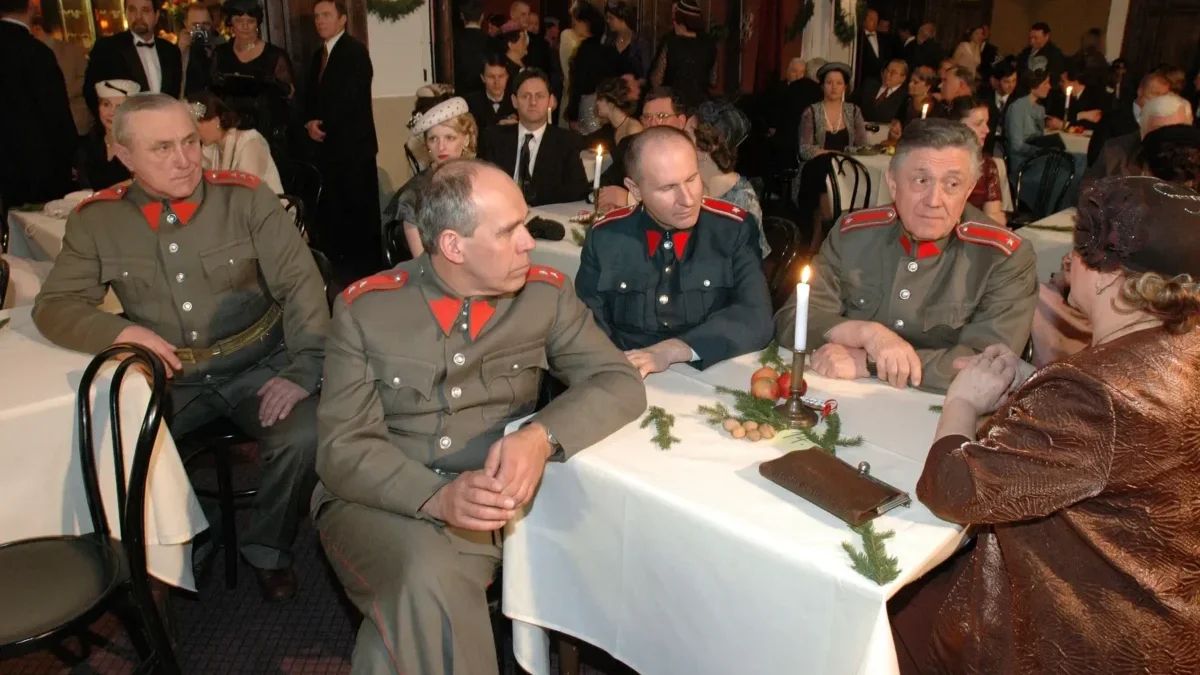 Four men in vintage gray military uniforms with red shoulder boards sit at a candlelit banquet table as guests in formal attire mingle in the background, festively decorated room behind them.