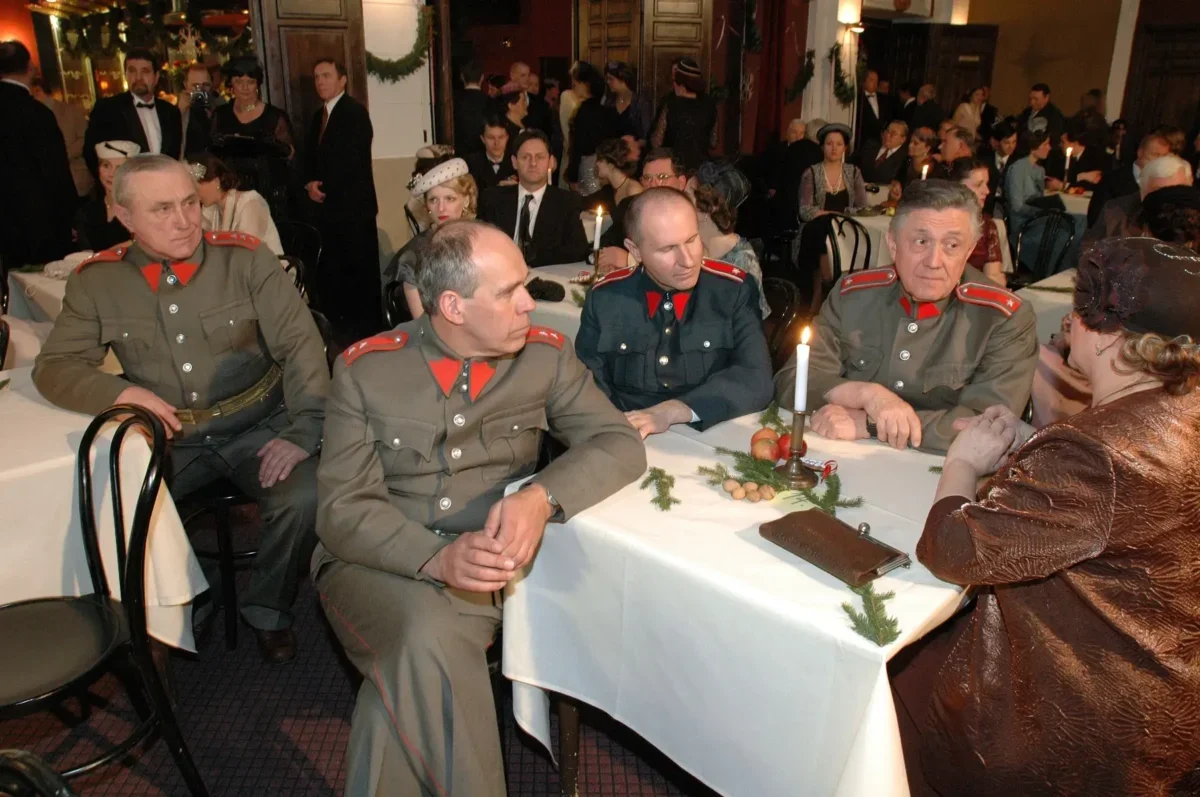 Four men in vintage gray military uniforms with red shoulder boards sit at a candlelit banquet table as guests in formal attire mingle in the background, festively decorated room behind them.