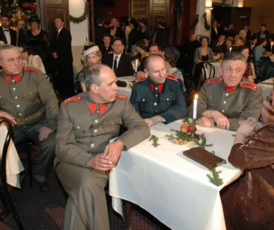 Four men in vintage gray military uniforms with red shoulder boards sit at a candlelit banquet table as guests in formal attire mingle in the background, festively decorated room behind them.