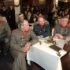 Four men in vintage gray military uniforms with red shoulder boards sit at a candlelit banquet table as guests in formal attire mingle in the background, festively decorated room behind them.