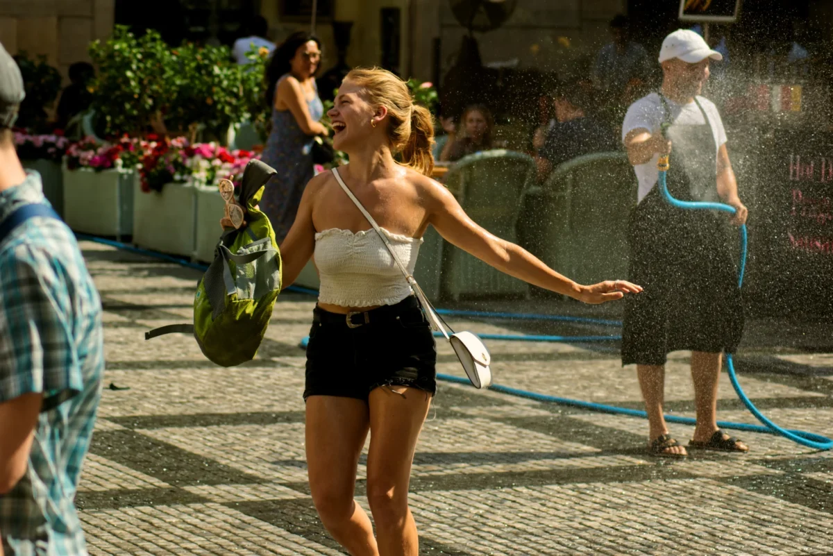 Woman in a white strapless top laughs with arms outstretched as water sprays from a hose at an outdoor event.