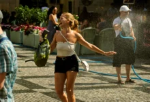 Woman in a white strapless top laughs with arms outstretched as water sprays from a hose at an outdoor event.
