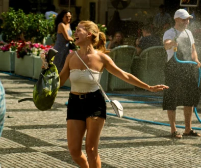 Woman in a white strapless top laughs with arms outstretched as water sprays from a hose at an outdoor event.
