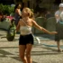 Woman in a white strapless top laughs with arms outstretched as water sprays from a hose at an outdoor event.