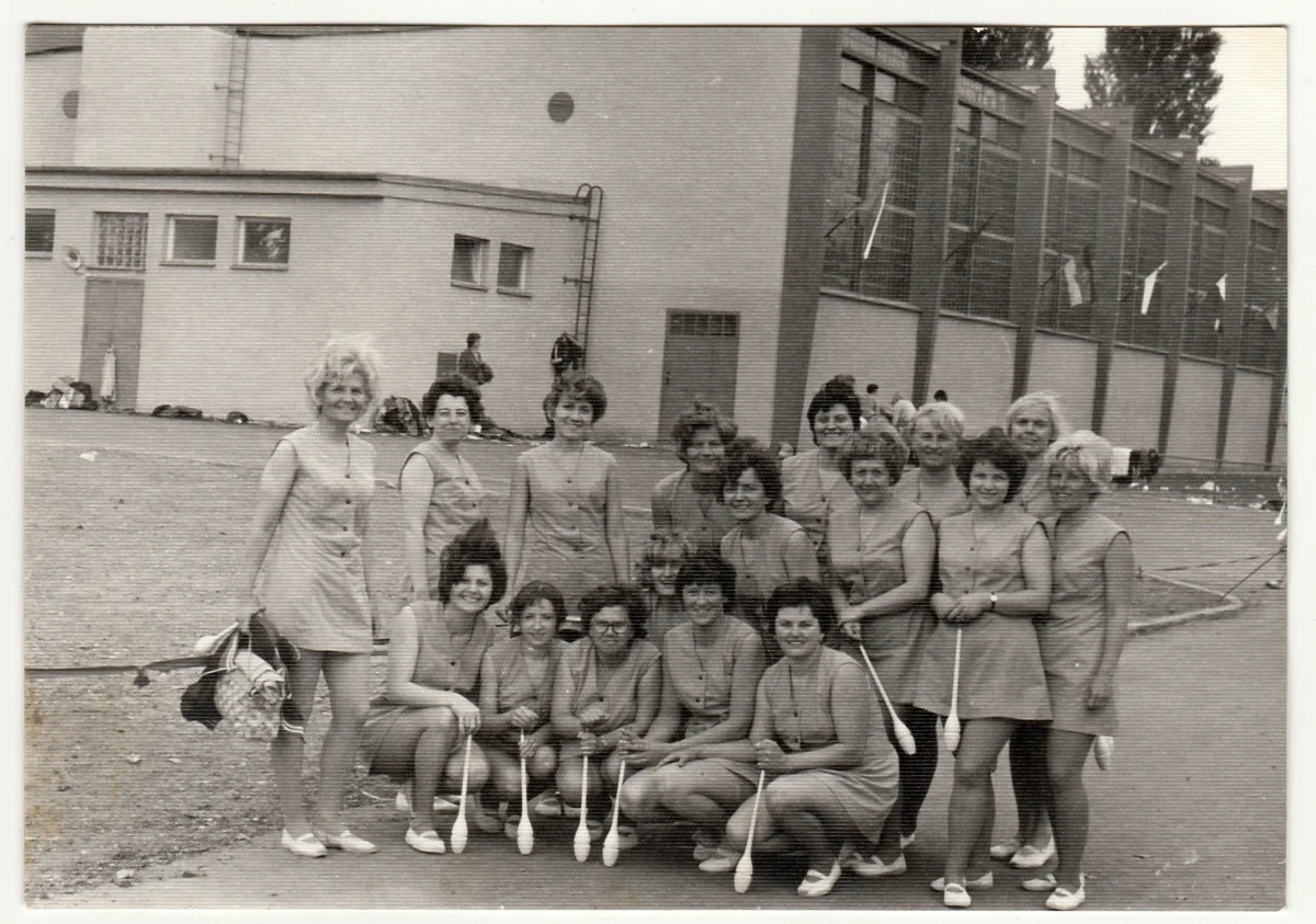 Group of women in swimsuits posing together outdoors, kneeling and standing, with a building behind them.