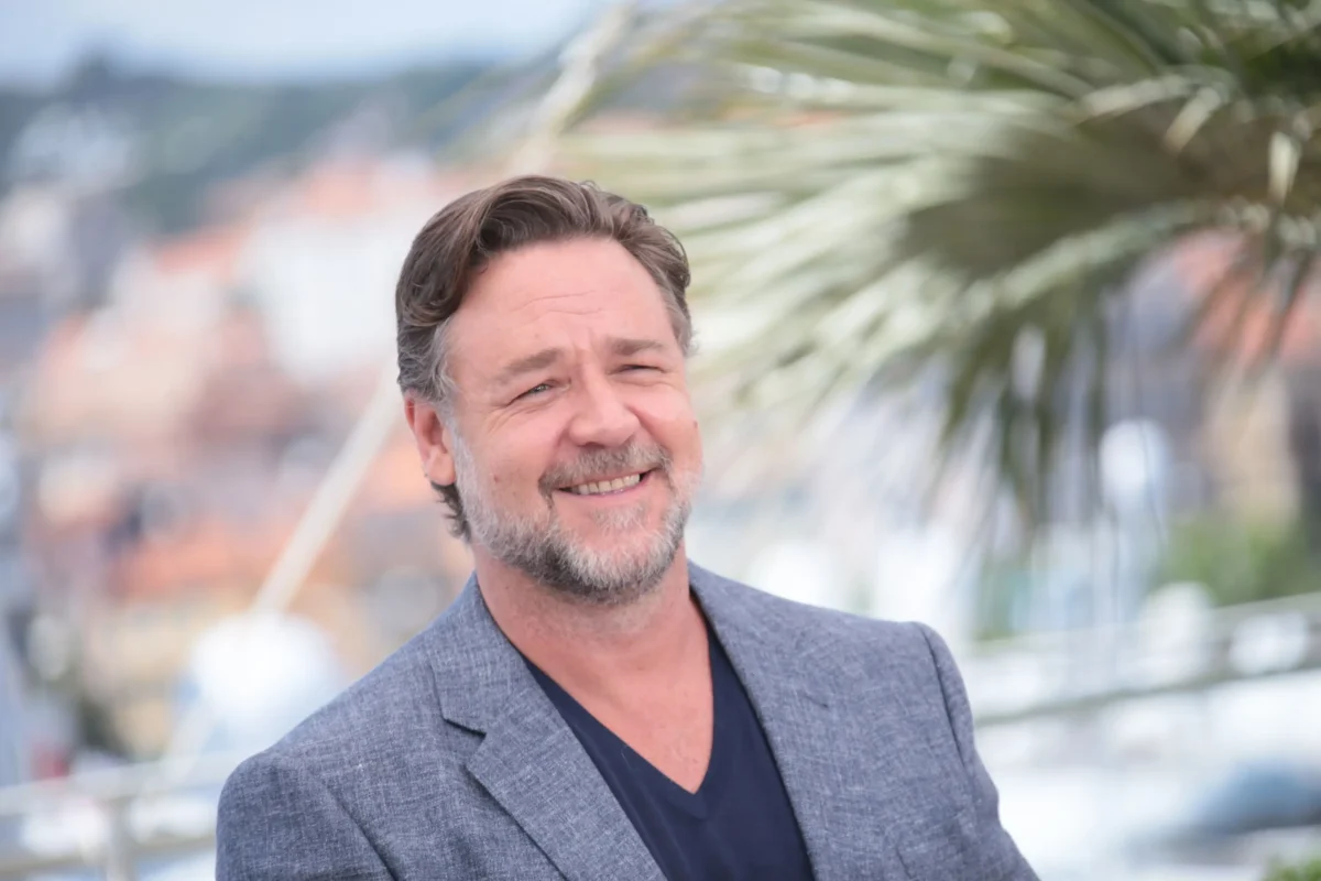 rusle | TVGURU.cz Russel Crowe v benátkách v šedivím oblek se usmívá a za ním je vidět palma