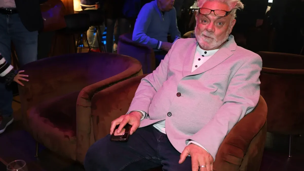 Older man with pink hair and glasses on his head sits in a pink blazer, holding a smartphone at a social event.