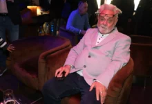 Older man with pink hair and glasses on his head sits in a pink blazer, holding a smartphone at a social event.