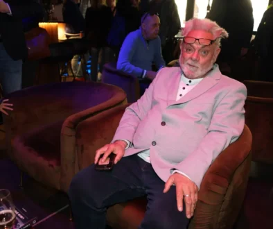 Older man with pink hair and glasses on his head sits in a pink blazer, holding a smartphone at a social event.