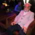 Older man with pink hair and glasses on his head sits in a pink blazer, holding a smartphone at a social event.