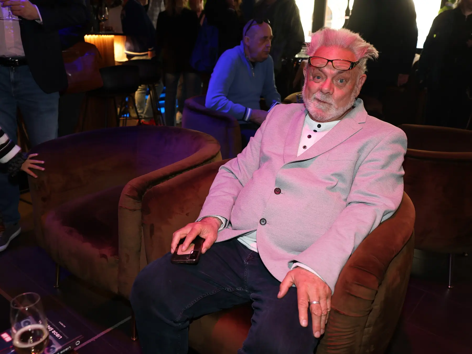 Older man with pink hair and glasses on his head sits in a pink blazer, holding a smartphone at a social event.