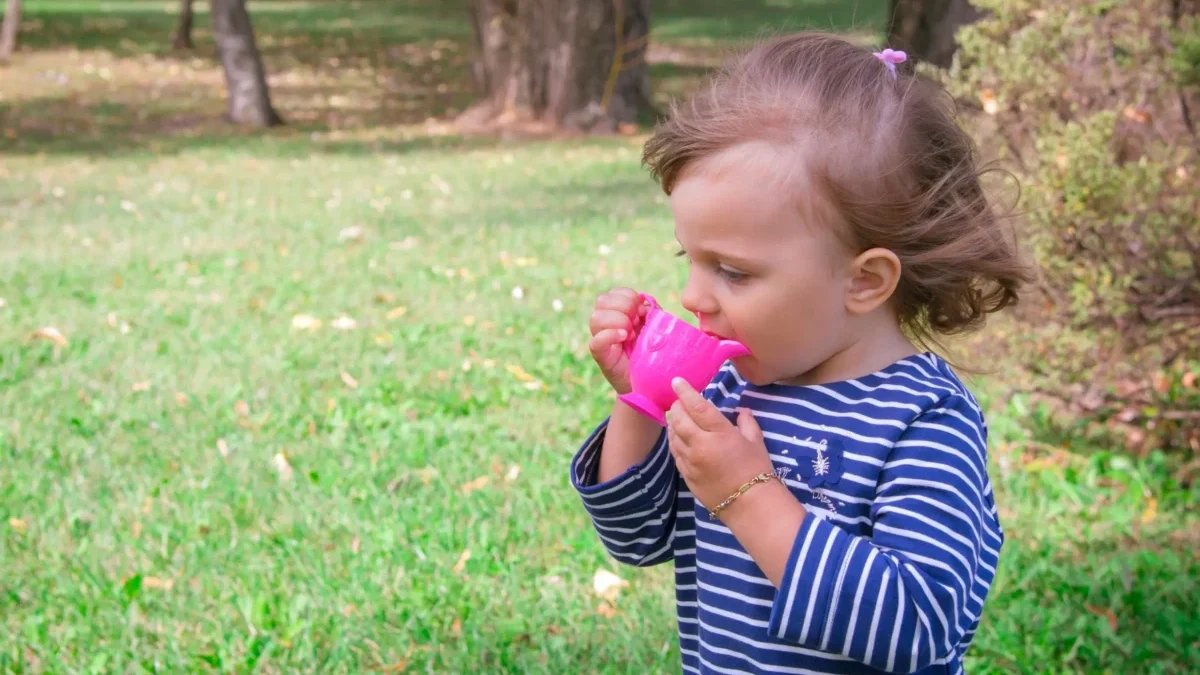 Young girl in a blue-and-white striped shirt drinks from a bright pink sippy cup in a park with green grass and trees in the background.