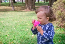 Young girl in a blue-and-white striped shirt drinks from a bright pink sippy cup in a park with green grass and trees in the background.