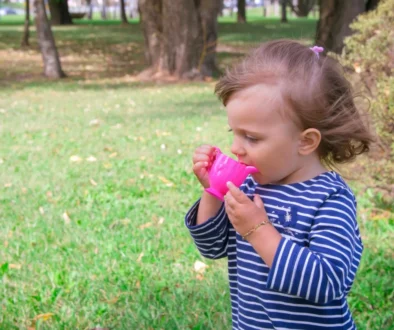 Young girl in a blue-and-white striped shirt drinks from a bright pink sippy cup in a park with green grass and trees in the background.