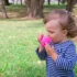 Young girl in a blue-and-white striped shirt drinks from a bright pink sippy cup in a park with green grass and trees in the background.