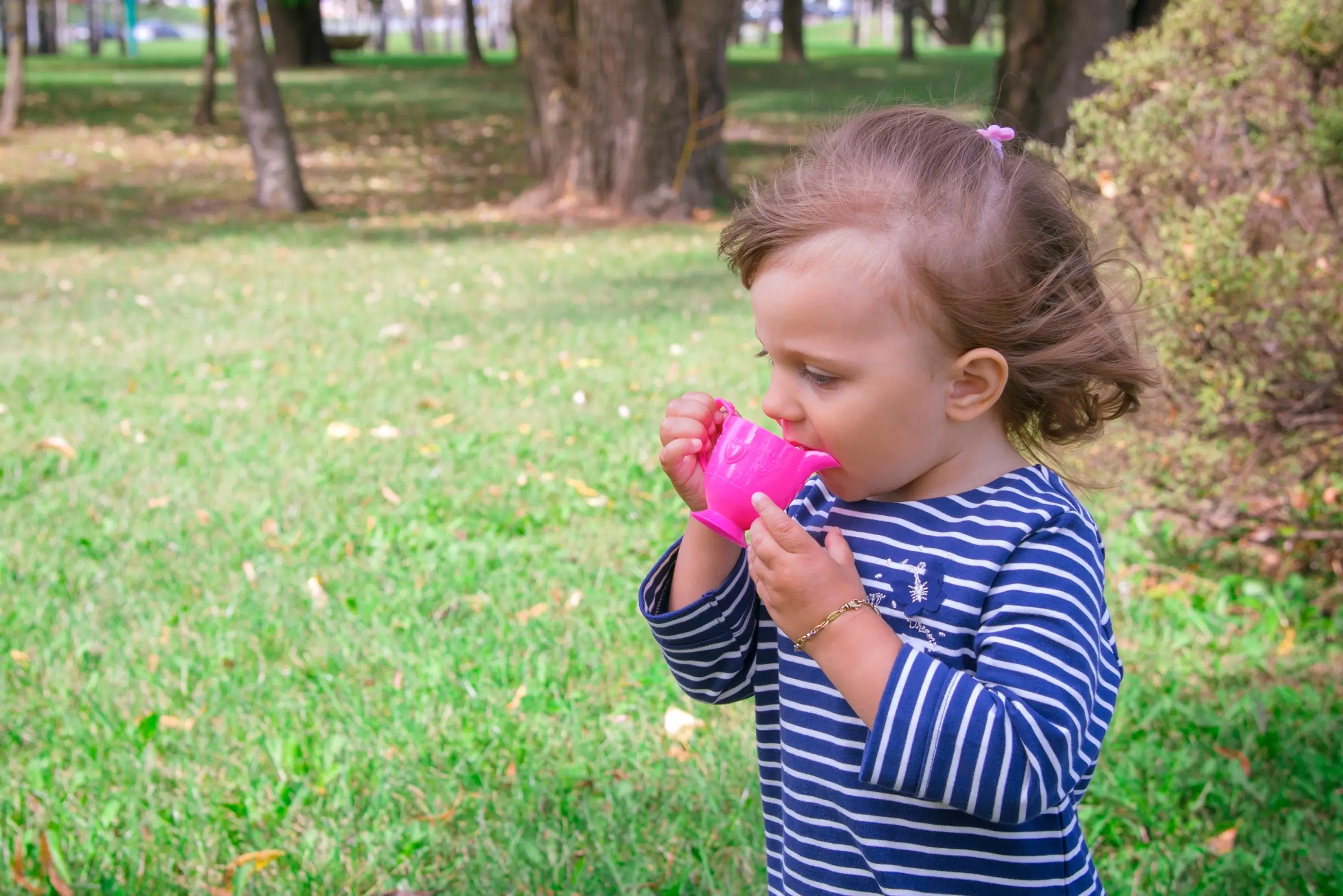 Young girl in a blue-and-white striped shirt drinks from a bright pink sippy cup in a park with green grass and trees in the background.