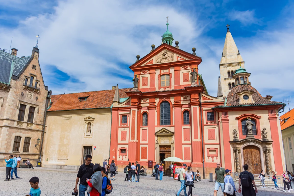 People walk and gather in a cobblestone square in front of a red ornate church facade with statues and a tower, under a blue sky.