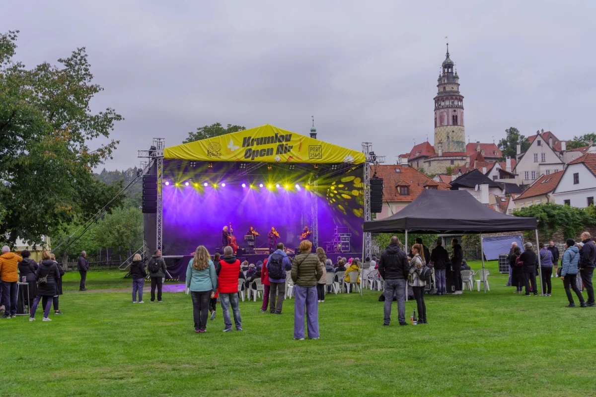Outdoor concert on a stage with purple lights, a yellow Krumlov Open Air banner, and an audience standing on the grassy square; historic town towers in the background.