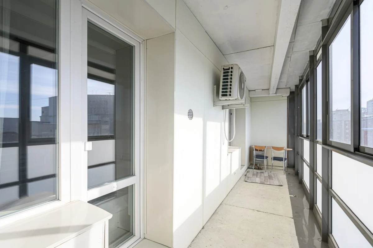 Narrow balcony along glass walls with a wall-mounted outdoor air conditioner and a small table with chairs in the far corner.