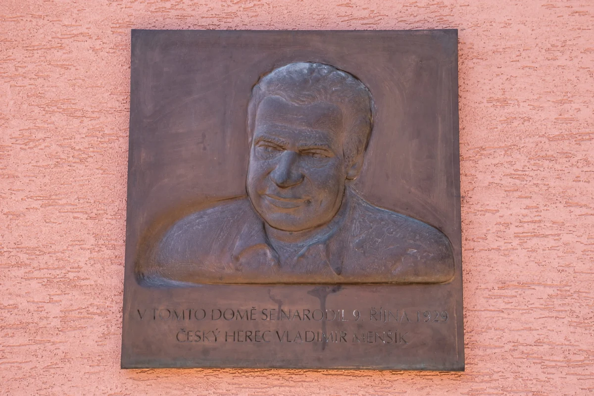Bronze relief plaque featuring a man’s head and shoulders on a dark panel mounted to a pink wall; Czech inscription at the bottom.