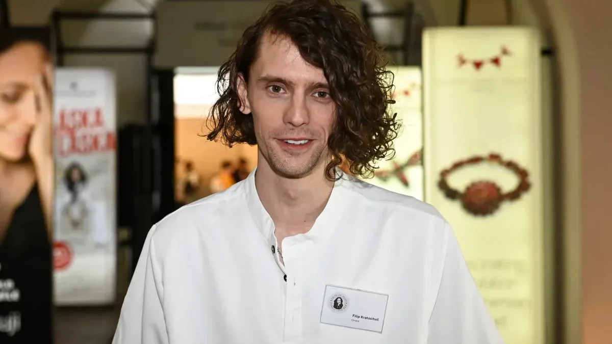 Man with shoulder-length wavy hair in a white shirt smiles at the camera at an indoor event venue, name badge visible on chest.