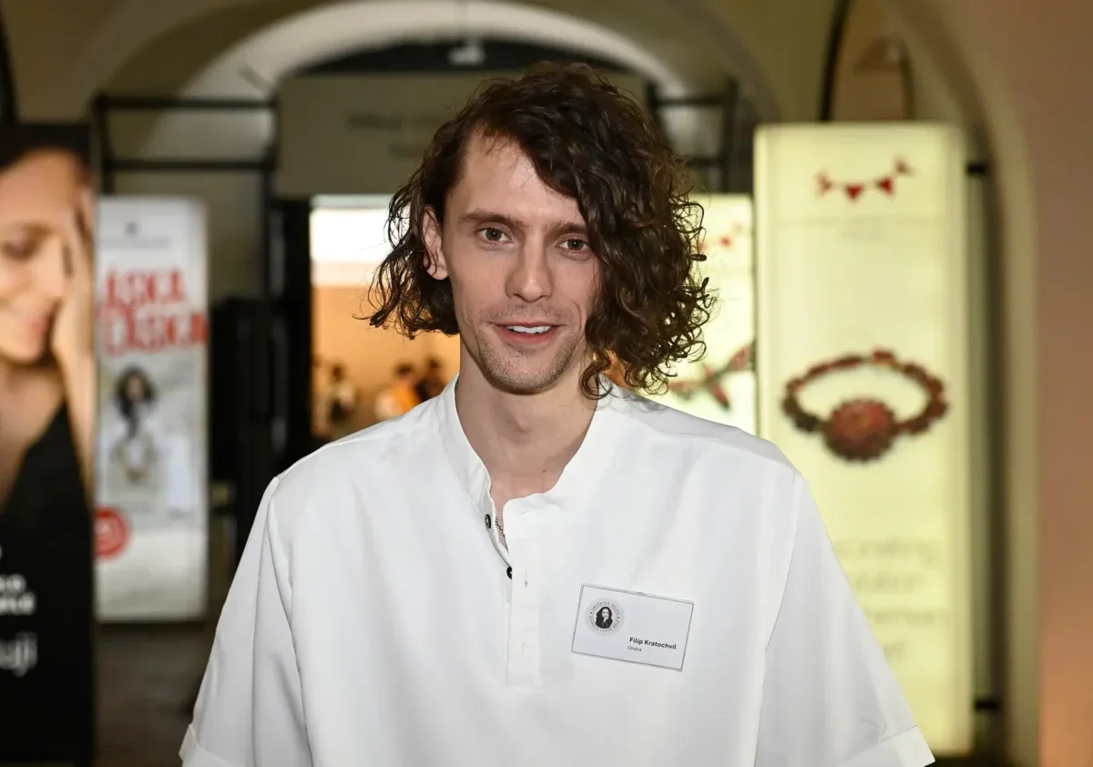 Man with shoulder-length wavy hair in a white shirt smiles at the camera at an indoor event venue, name badge visible on chest.