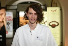 Man with shoulder-length wavy hair in a white shirt smiles at the camera at an indoor event venue, name badge visible on chest.