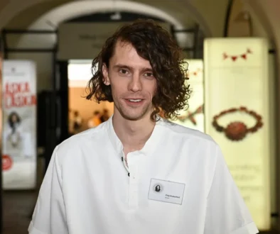 Man with shoulder-length wavy hair in a white shirt smiles at the camera at an indoor event venue, name badge visible on chest.