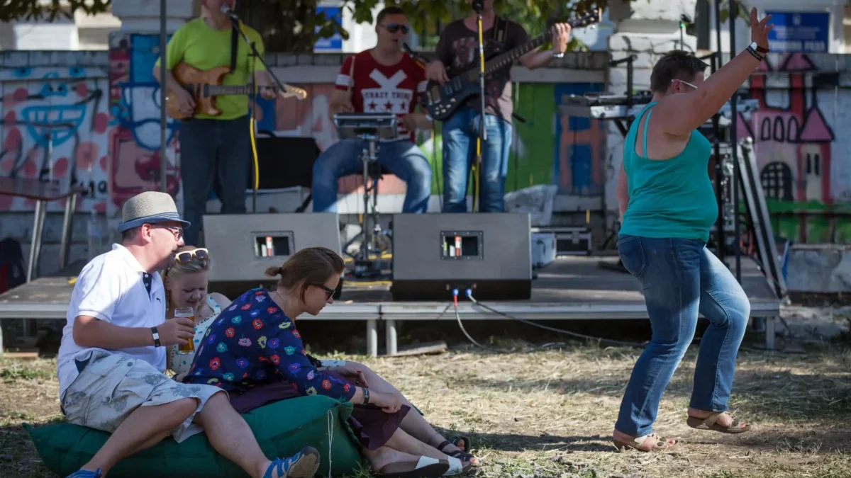 A small outdoor concert with a band on a graffiti-covered stage; seated audience on a blanket in the foreground, including a man in a hat and two women enjoying drinks.