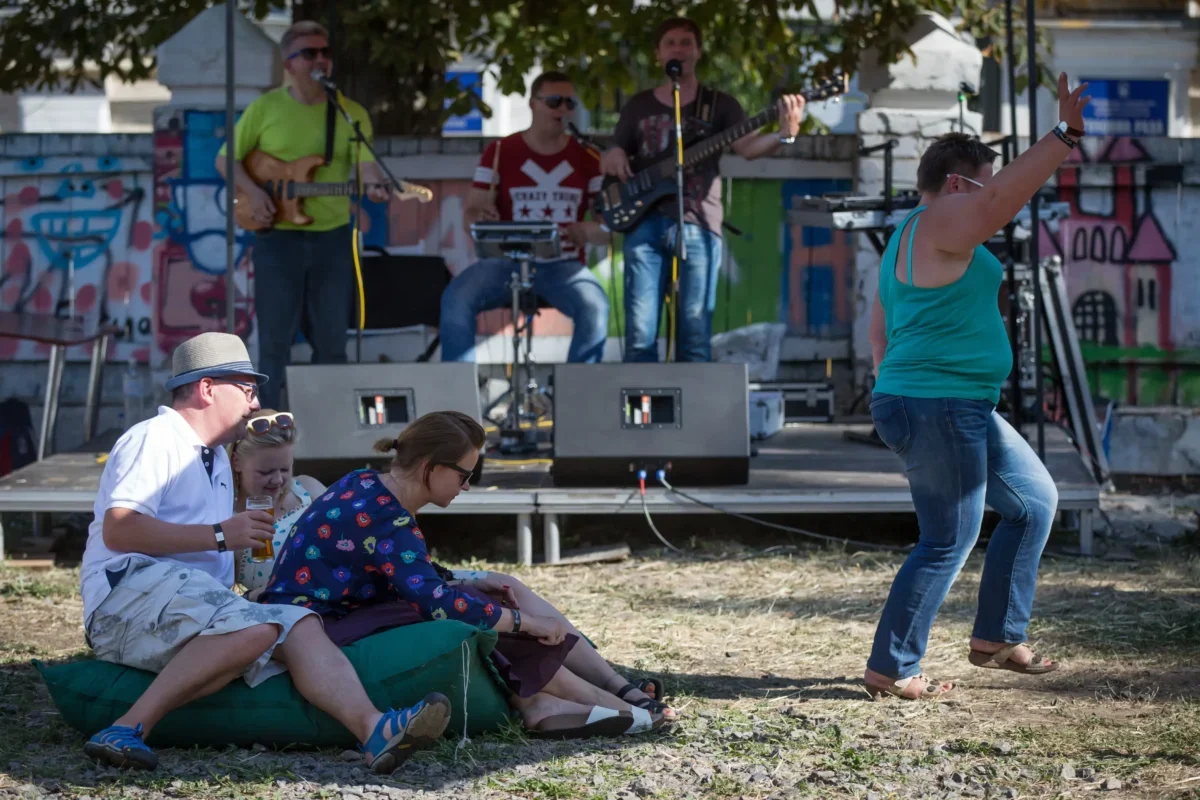 A small outdoor concert with a band on a graffiti-covered stage; seated audience on a blanket in the foreground, including a man in a hat and two women enjoying drinks.