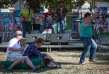 A small outdoor concert with a band on a graffiti-covered stage; seated audience on a blanket in the foreground, including a man in a hat and two women enjoying drinks.