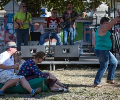 A small outdoor concert with a band on a graffiti-covered stage; seated audience on a blanket in the foreground, including a man in a hat and two women enjoying drinks.