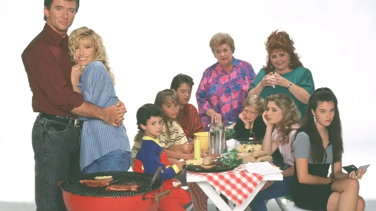 A family crowd around a picnic table at a barbecue, with a couple embracing on the left and others watching or chatting nearby.