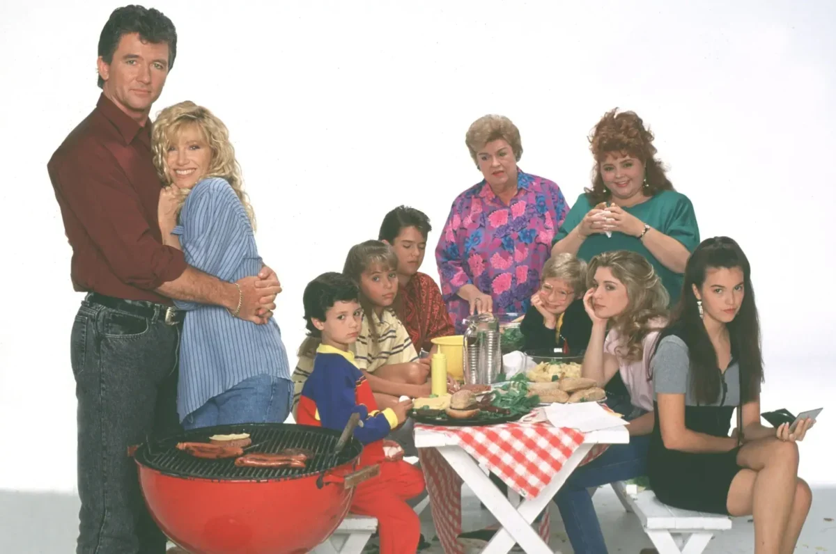 A family crowd around a picnic table at a barbecue, with a couple embracing on the left and others watching or chatting nearby.