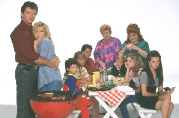 A family crowd around a picnic table at a barbecue, with a couple embracing on the left and others watching or chatting nearby.