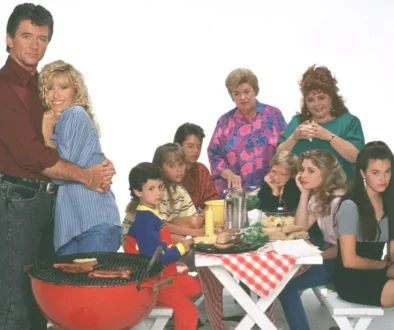 A family crowd around a picnic table at a barbecue, with a couple embracing on the left and others watching or chatting nearby.
