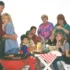 A family crowd around a picnic table at a barbecue, with a couple embracing on the left and others watching or chatting nearby.