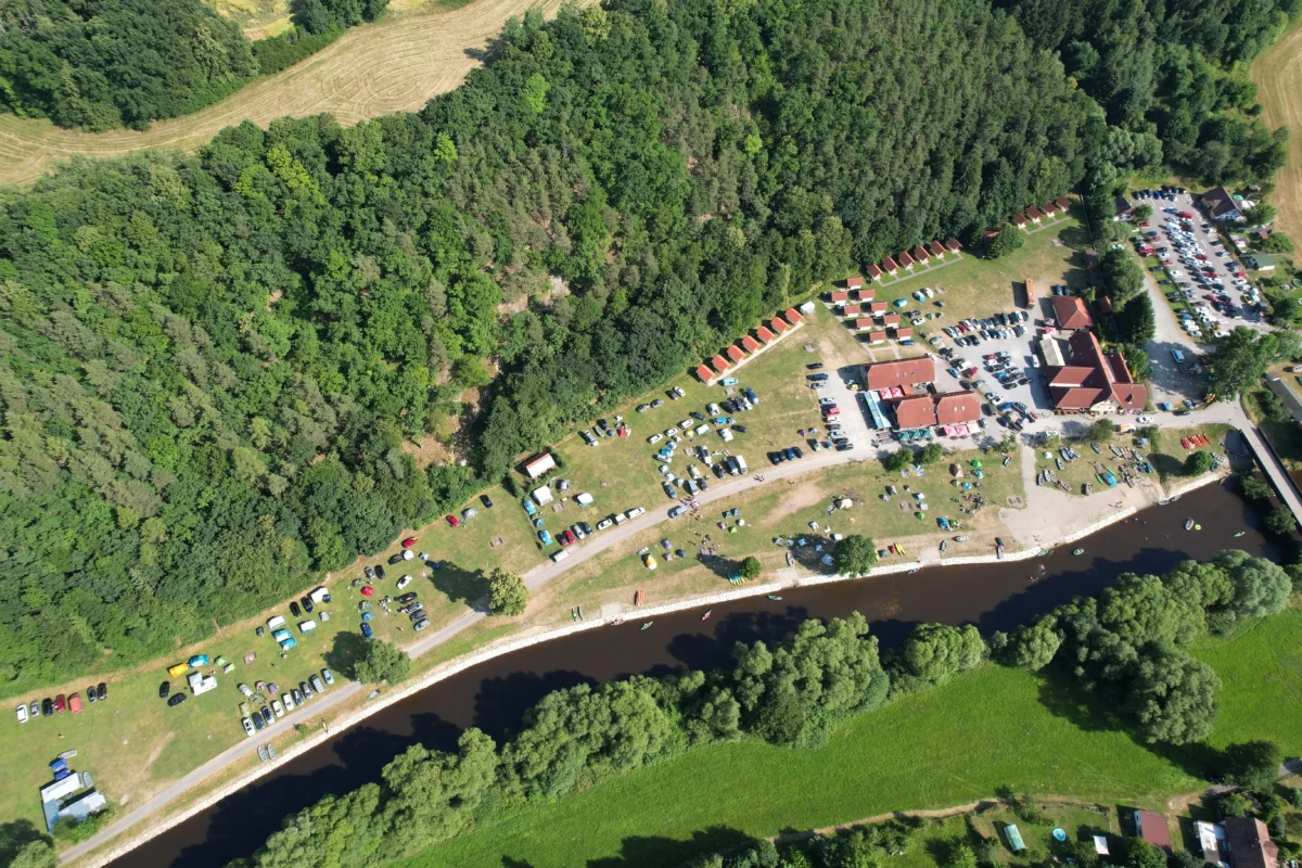 Aerial view of a campground along a river with rows of parked cars, small cabins, and a dense tree line nearby.
