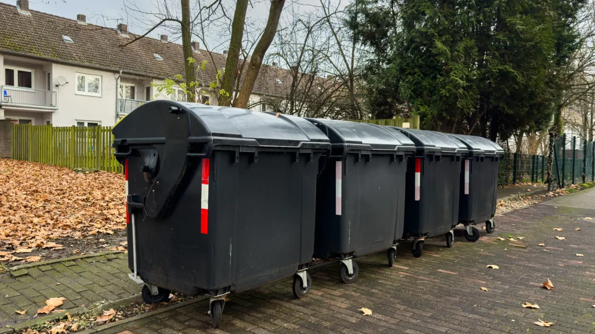 Row of large black dumpsters on a residential street with fallen leaves and apartment buildings in the background.