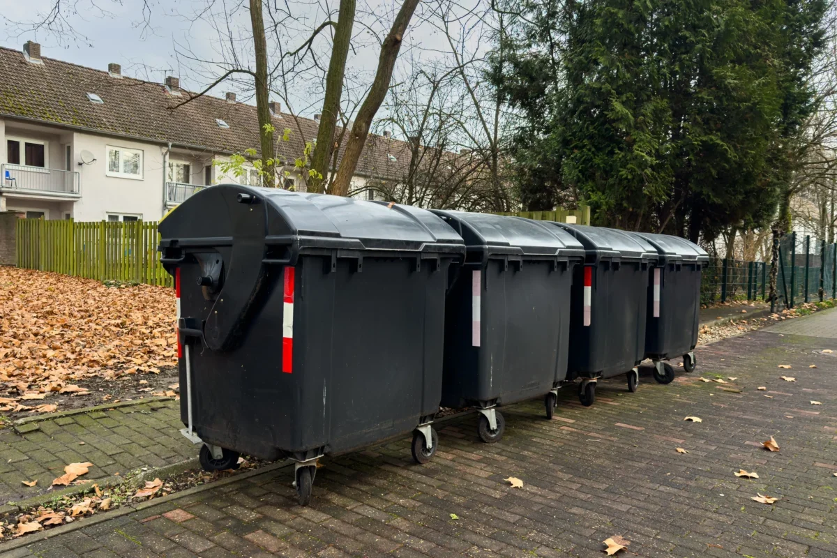 Row of large black dumpsters on a residential street with fallen leaves and apartment buildings in the background.