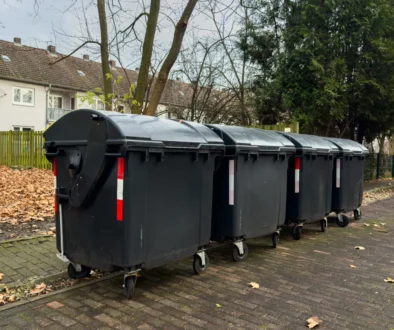 Row of large black dumpsters on a residential street with fallen leaves and apartment buildings in the background.