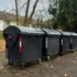 Row of large black dumpsters on a residential street with fallen leaves and apartment buildings in the background.