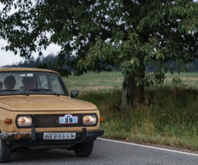 Old beige vintage car on a country road with a large tree in the background; three people visible through the windshield.